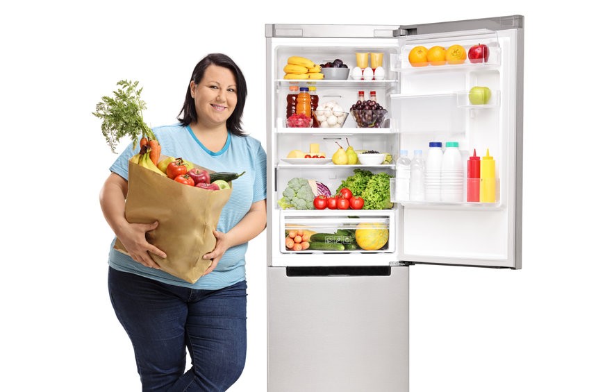 אישה שמנה מחזיקה שקית עם ירקות, עומדת ליד מקרר